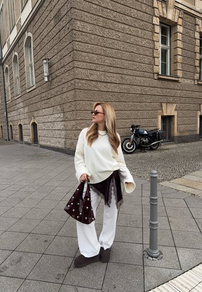Suéter blanco de gran tamaño, pantalones blancos de pierna ancha, botas marrón oscuro y un bolso burdeos brillante con detalles de perlas. Fondo de edificio texturizado.