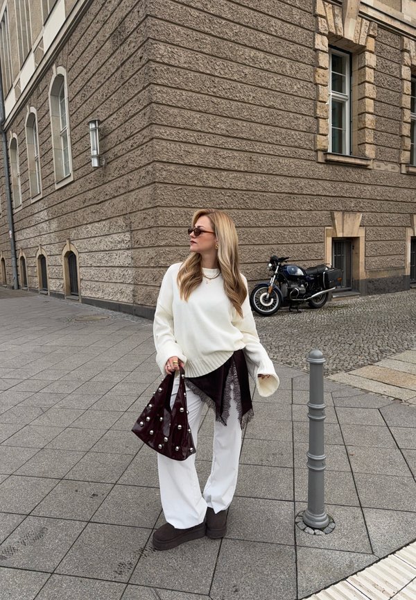 Pulover alb supradimensionat, pantaloni albi largi, cizme maro închis și o geantă lucioasă burgund cu accente de perle. Fundal cu clădire texturată.