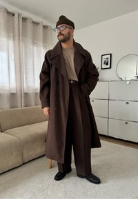 Mężczyzna w ciemnobrązowym płaszczu, brązowych spodniach, beżowym swetrze, czapce i okularach przeciwsłonecznych stojący w nowoczesnym salonie z beżową sofą i białą szafką.