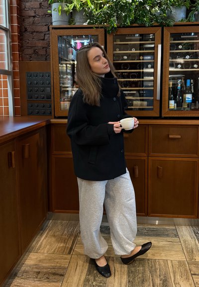 Mujer con abrigo negro y pantalones grises sosteniendo una taza blanca, de pie en una cocina de madera con armarios para vino y plantas encima.