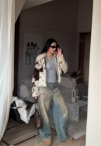 Jakna iz umetnega krzna z rjavimi in belimi madeži, siva majica, prevelike pošivane modre kavbojke, rjavi mule čevlji in temna torbica. Minimalističen notranji prostor.