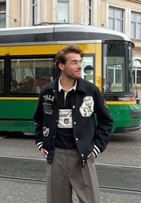 Veste universitaire noire avec des rayures blanches, broderie Yale et écusson de chien, portée sur un polo noir et blanc, avec un pantalon beige. Bus en arrière-plan.