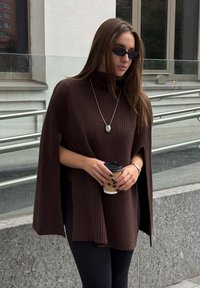 Poncho a coste marrone con colletto alto e aperture laterali, abbinato a leggings neri. Tiene in mano una tazza di caffè e indossa una collana d'argento elegante.
