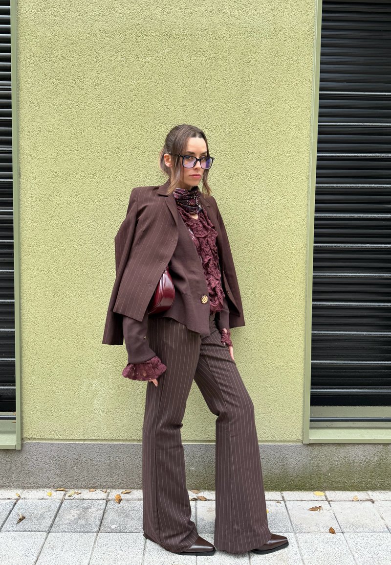 Frau in braunem Nadelstreifenanzug mit Rüschenbluse, Schal und Brille steht vor einer hellgrünen Wand auf einem gepflasterten Bürgersteig.