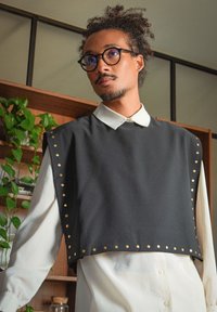 Svart kort topp med guldstift längs kanterna, lager på en vit skjorta med krage. Slät, strukturerad material med en modern design.