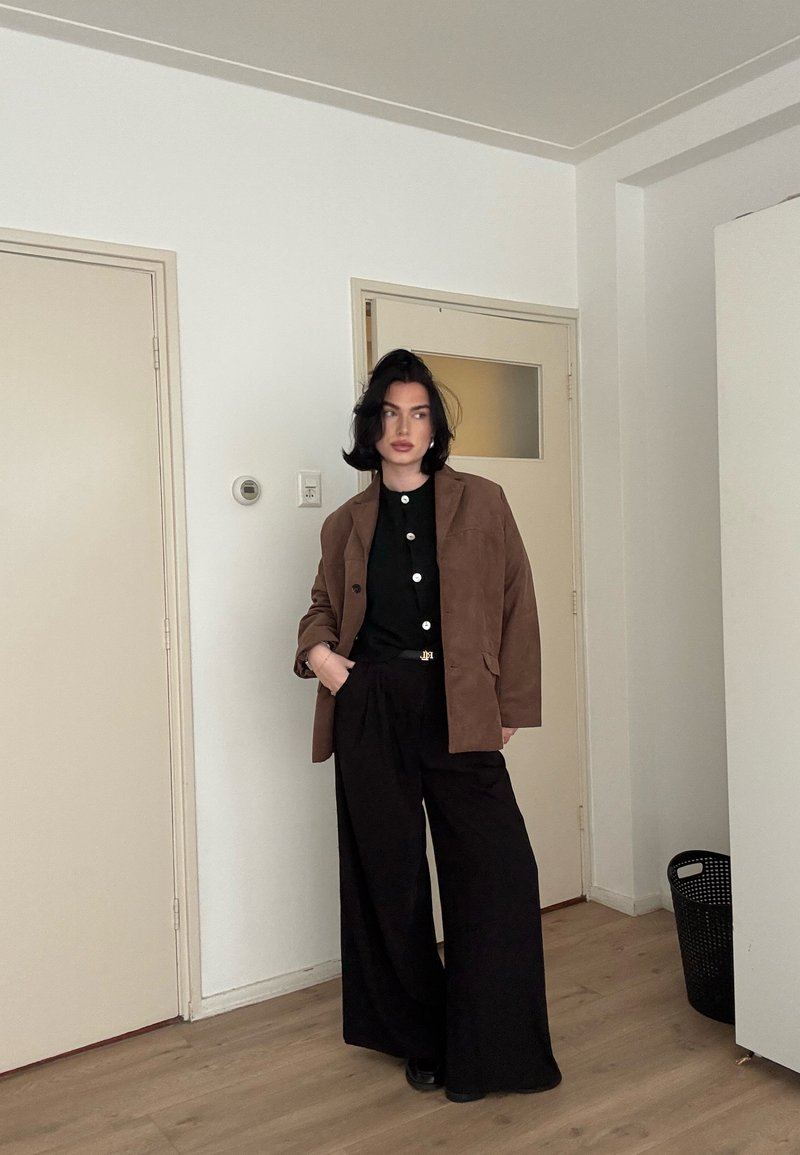 Femme aux cheveux châtains mi-longs portant une veste marron sur un haut noir et un pantalon noir à jambes larges, debout à l'intérieur près de portes fermées.