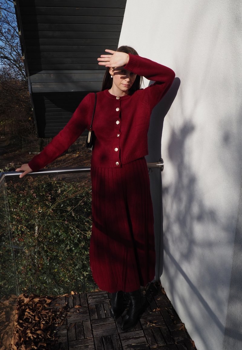 Cardigan lavorato a maglia color bordeaux con dettagli a bottoni, abbinato a una gonna lunga a costine dello stesso colore. La texture liscia e il design aderente accentuano la silhouette.
