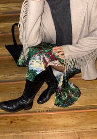 Άτομο καθισμένο σε ξύλινα σκαλιά, φορώντας μπεζ παλτό με φούντες, πολύχρωμη φούστα με σχέδια, μαύρες μπότες μέχρι το γόνατο και με μαύρη τσάντα στο πλάι.