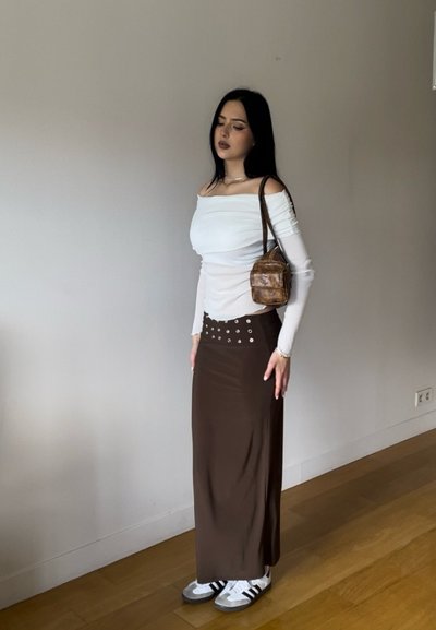 Top blanco sin mangas con hombros al descubierto, falda maxi marrón con tachuelas plateadas y zapatillas grises de Adidas con rayas negras. Bolso cruzado marrón.