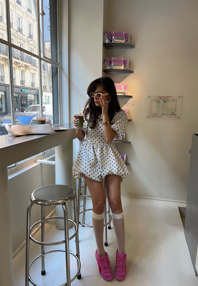 Vestido blanco con lunares y mangas abullonadas, combinado con calcetas blancas hasta la rodilla y zapatillas rosas. Bebida verde en mano, sentado en una barra.