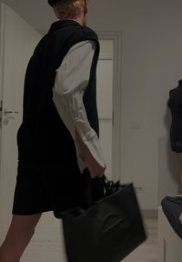 Saco tote preto com alças superiores e um detalhe circular. Modelo vestindo um top de malha preto sem mangas sobre uma camisa de manga longa branca e calções pretos.