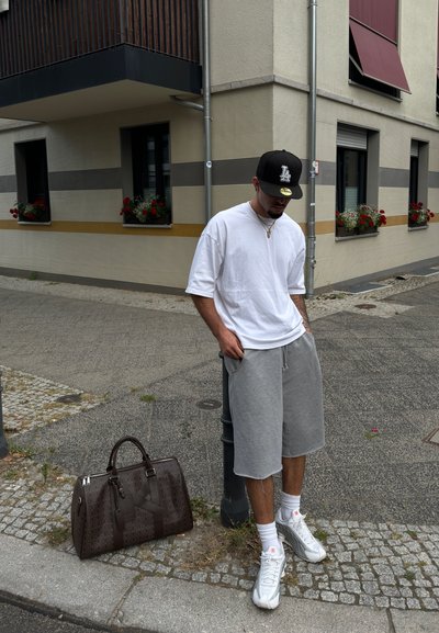 Pantalones cortos grises, camiseta blanca oversized, gorra negra y zapatillas blancas. Bolsa de viaje de patrón marrón en el suelo junto a una farola.