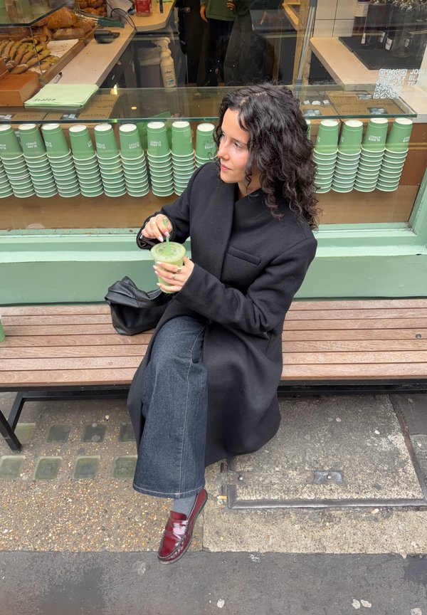 Žena s kučeravými vlasmi v čiernom kabáte sedí na lavičke pred kaviarňou, drží a mieša zelené smoothie so slamkou.