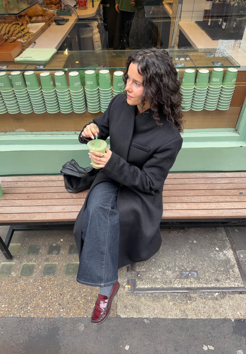 Donna con capelli ricci e cappotto nero seduta su una panchina fuori da un caffè, che tiene e mescola un frullato verde con una cannuccia.