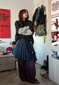 En modell bär en svart oversized sweatshirt med grå ärmar, en blå rutigt kjol och lösa lila byxor. Bakgrunden har olika väskor och affischer.