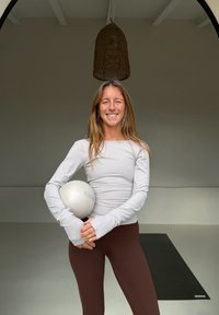 Femme en tenue de sport tenant un ballon de Pilates, debout sur un sol de gym avec un tapis de yoga noir derrière elle et un luminaire en osier au-dessus.