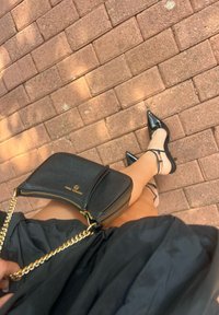 Μαύρη δερμάτινη τσάντα με χρυσή αλυσίδα, κλείσιμο με φερμουάρ και λεπτομέρεια λογότυπου; μαύρες λαμπερές δερμάτινες γόβες με λουράκι στον αστράγαλο σε επιφάνεια τούβλου.