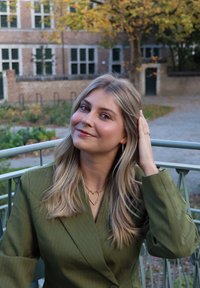 Olivengrønn blazer med tynne striper og V-hals, kombinert med et hjerteformet gullkjede. Mykt bølget blondt hår og en nøytral bakgrunn.