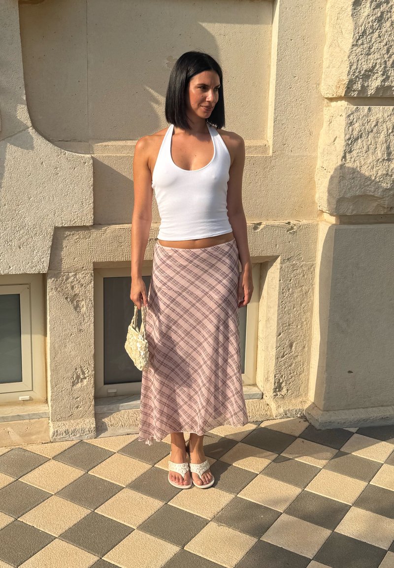 Γυναίκα με ίσια μαύρα μαλλιά που φοράει λευκή αμάνικη μπλούζα, ροζ καρό φούστα, λευκές σανδάλες και κρατά μια μικρή λευκή τσάντα έξω.