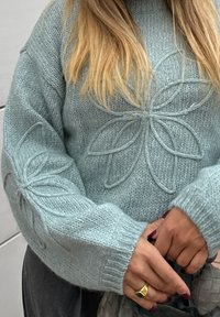 Vaaleansininen neulepusero, jossa on kukkakuosi, paksu rakenne ja joustinvärjätyt hihansuut. Kädet ristissä vyötäröllä, sormessa kultasormus.