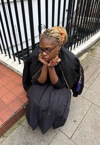 Persona sēž uz ietves, valkājot melnu bomberu jakas un garu melnu kleitu ar teksturētu audumu, zelta rotaslietas un pinumus ar oranžu matu gumiju.