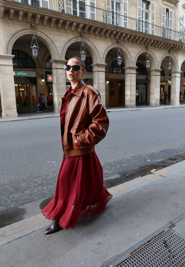 Une femme portant des lunettes de soleil, une veste en cuir marron et une robe rouge fluide passe devant une arcade en pierre dans une rue de la ville.