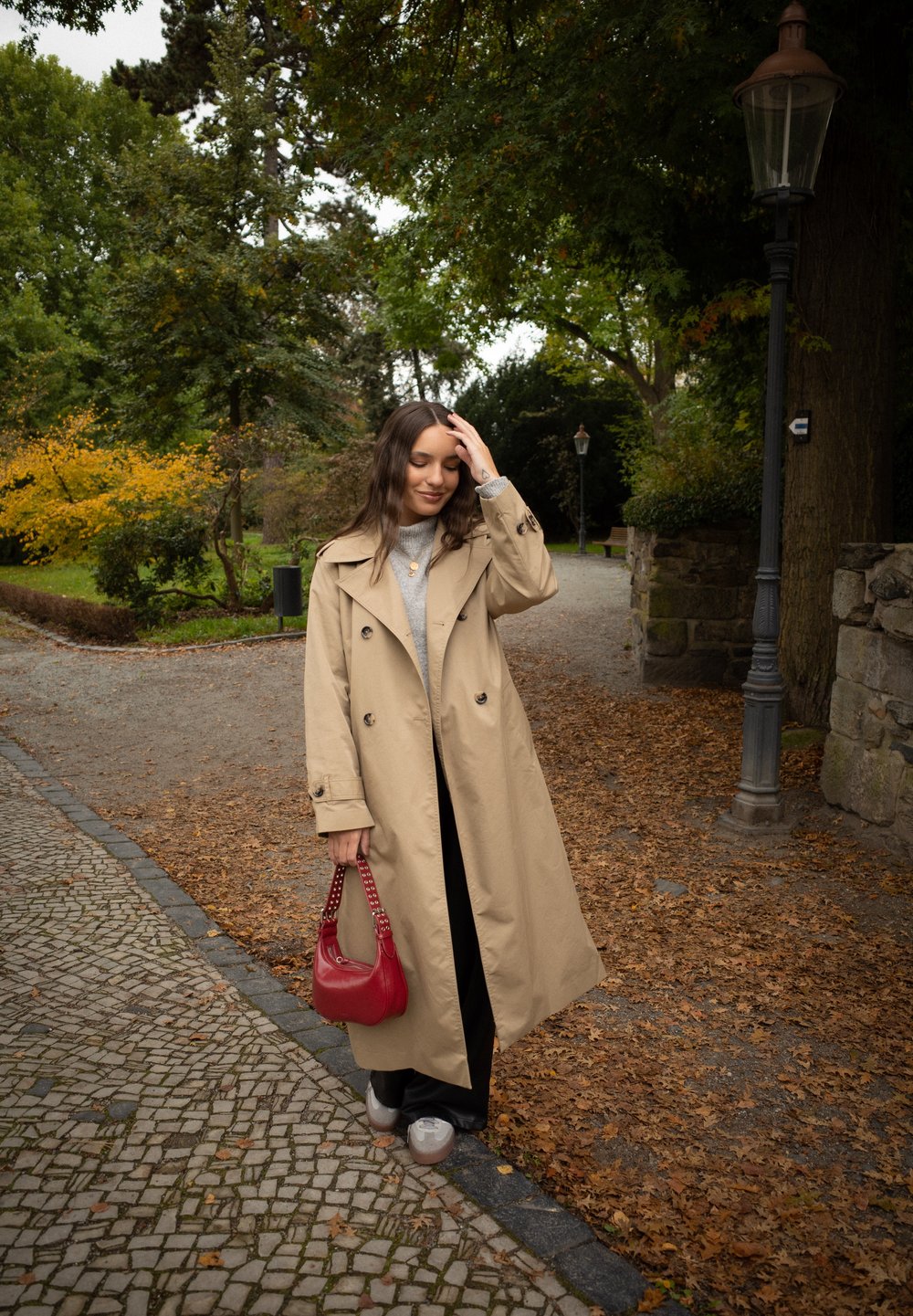Trench coat bege com botões duplos, combinado com um suéter cinza, calças pretas e uma bolsa vermelha. Fundo texturizado de folhas de outono.