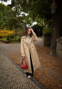 Trench coat bege com botões duplos, combinado com um suéter cinza, calças pretas e uma bolsa vermelha. Fundo texturizado de folhas de outono.