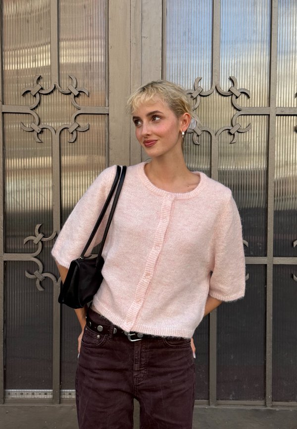 Maglione corto maniche a sbuffo color rosa chiaro con scollatura rotonda e chiusura a bottoni, abbinato a pantaloni marrone scuro e una borsa a tracolla nera.