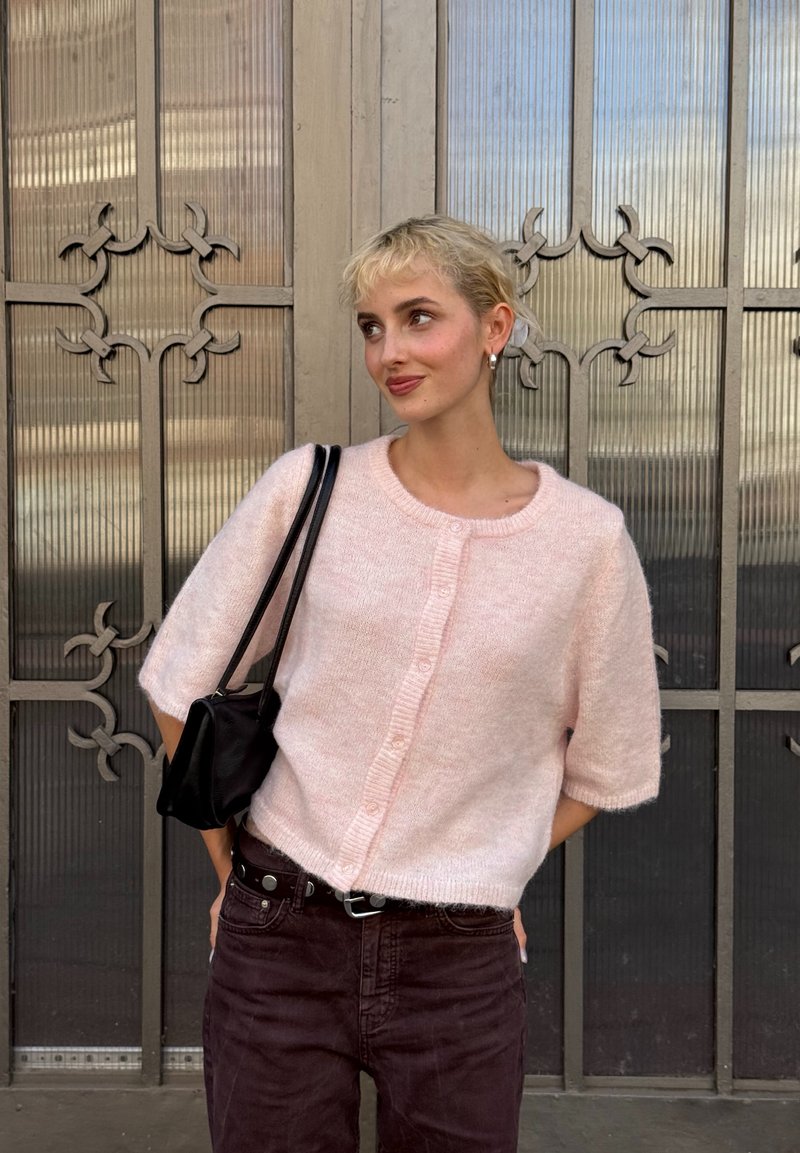 Heller rosa, kurzärmliger Pullover mit Rundhalsausschnitt und Knopfschluss, kombiniert mit dunkellbraunen Hosen und einer schwarzen Schultertasche.