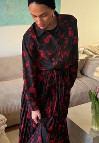 Naine reguleerimas musta ja punase lillemustrilise volditud kleiti, seistes elutoas beež diivani ja laual asuva tulpidevaasiga.