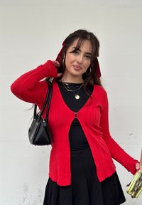 Jeune femme aux cheveux foncés attachés avec des rubans rouges, portant un cardigan rouge sur un haut et une jupe noirs, tenant un sac à main noir et un bouquet d'asperges.