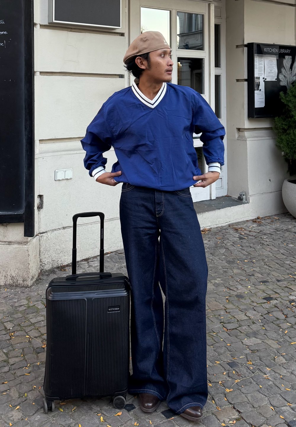 Maillot bleu à manches longues avec des accents blancs, associé à un jean denim foncé à jambes larges. Valise noire avec un design texturé et roues.