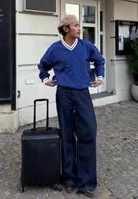Maillot bleu à manches longues avec des accents blancs, associé à un jean denim foncé à jambes larges. Valise noire avec un design texturé et roues.