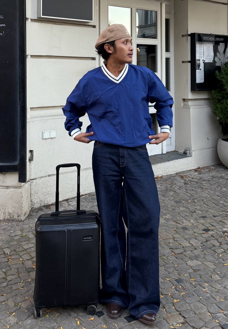 Maillot bleu à manches longues avec des accents blancs, associé à un jean en denim foncé à jambes larges. Valise noire avec design texturé et roues.