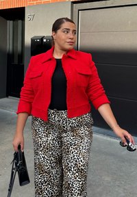 Rød tekstureret jakke med to for-tryk og parret med brede leopardmønstrede bukser. Holder en sort håndtaske og solbriller. Ensfarvet baggrund.