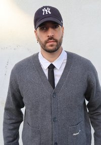 Cardigan gris en laine avec un col en V, deux poches avant et des boutons foncés, porté sur une chemise blanche et une cravate noire. Casquette de baseball noire sur la tête.