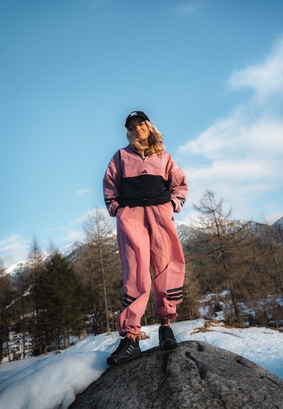 Chándal de Adidas en rosa y negro con chaqueta cortaviento y pantalones ajustados. Gorra negra, zapatos negros, de pie sobre una roca con un paisaje nevado de fondo.