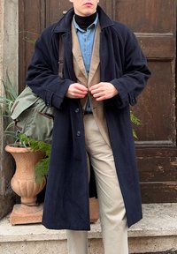 Jeune personne vêtue en couches superposées comprenant un manteau bleu marine, un blazer marron, une chemise en jean et un pantalon beige, en train de fermer les boutons du manteau devant une porte en bois.