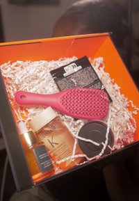 Różowa szczotka do włosów z teksturowanym uchwytem, dwa produkty do włosów w przezroczystych i czarnych pojemnikach oraz błyszczący olejek, zaprezentowane w pomarańczowym pudełku z pociętą na wiór papierową wyściółką.