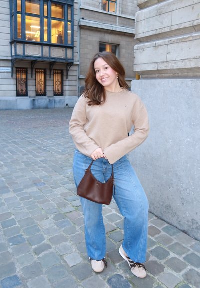 Joven sonriente, de pie en una calle empedrada, viste un suéter beige, jeans azules, zapatillas blancas y sostiene un bolso marrón con ambas manos.