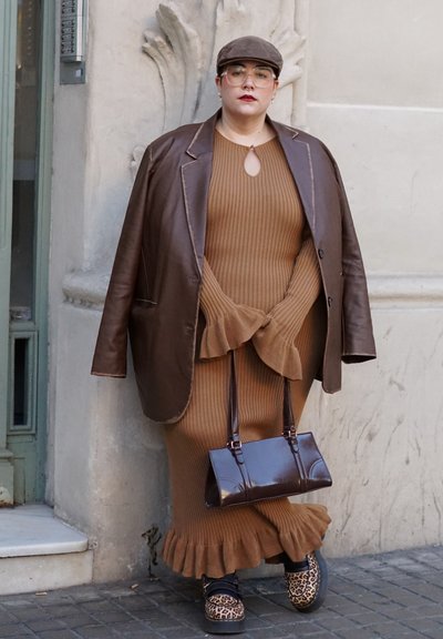 Persona vistiendo un vestido marrón de canalé con mangas y dobladillo con volantes, chaqueta de cuero marrón oversize, zapatos con estampado de leopardo, gorra, gafas y un bolso marrón.