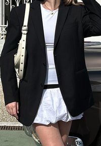 Blazer nero sartoriale su un top bianco aderente, abbinato a una gonna bianca a strati. Presenta una tasca singola e chiusura con bottone.