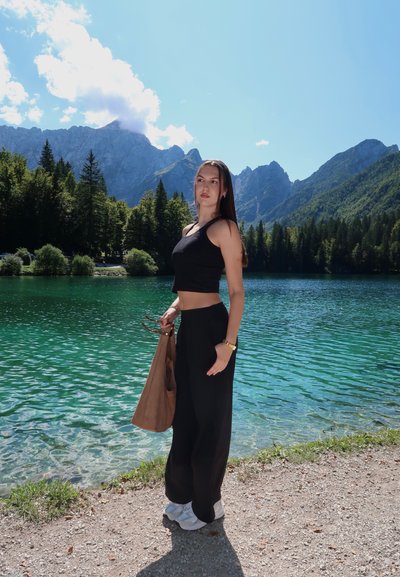 Top negro corto y pantalones anchos negros, combinados con zapatos blancos y un bolso marrón, están al lado de un lago turquesa con montañas al fondo.