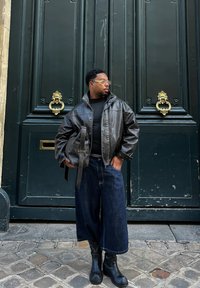 Jachetă din piele neagră, cămașă neagră, pantaloni cropped din denim închis, cizme negre, ținând o geantă maro, pe fondul unei uși verzi cu accesorii aurii.