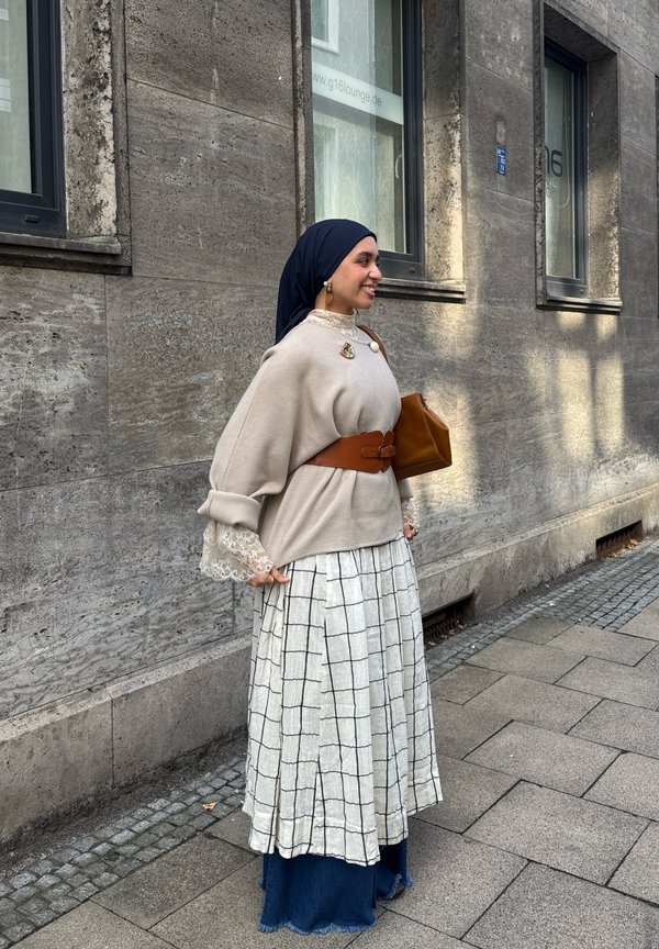 Nő bézs felsővel, kockás szoknyával, sötétkék hijabbal és barna övvel, aki egy kőépület mellett áll a városi járdán.