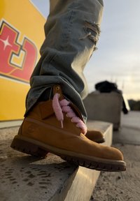 Gros plan sur une botte beige avec des lacets roses pelucheux, portée avec un jean bleu déchiré, reposant sur un rebord en béton à côté d'un mur jaune avec des lettres rouges.