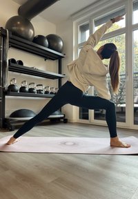 Személy, aki egy jógapózt végez világos rózsaszín jóga szőnyegen. Világos bézs kapucnis felsőt és fekete leggingset visel. Fitness felszerelések vannak a háttérben.