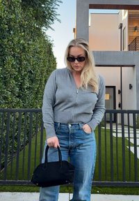 Femme aux longs cheveux blonds portant des lunettes de soleil, un pull gris à fermeture éclair et un jean bleu, tenant un sac à main noir, debout près d'une maison moderne et d'une haie.