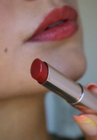 Rossetto rosso con una punta liscia e rotonda tenuto da una mano. Il bullet è in una custodia di metallo argentato con una finitura lucida.